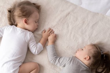 Çocukluk, uyku, rahatlama, aile, yaşam tarzı konsepti - 2 ve 3 yaşlarındaki iki genç, beyaz ve bej giysiler içinde öğle yemeğinde el ele tutuşarak bej ve beyaz bir yatakta uyurlar..