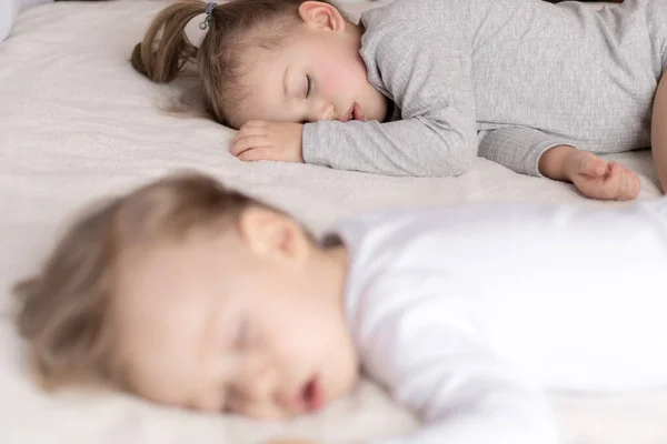 Infancia, sueño, relajación, familia, concepto de estilo de vida: dos niños pequeños de 2 y 3 años vestidos con traje de cuerpo blanco y beige duermen en una cama beige y blanca en el almuerzo tomados de la mano vista superior . — Foto de Stock