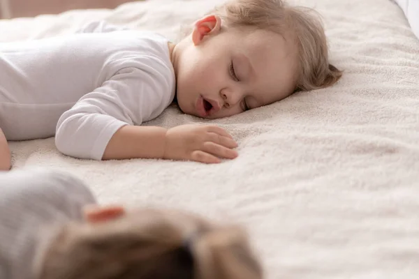 Infancia, sueño, relajación, familia, concepto de estilo de vida: dos niños pequeños de 2 y 3 años vestidos con traje de cuerpo blanco y beige duermen en una cama beige y blanca en el almuerzo tomados de la mano vista superior . — Foto de Stock