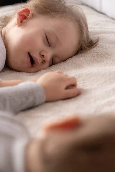 Infancia, sueño, relajación, familia, concepto de estilo de vida: dos niños pequeños de 2 y 3 años vestidos con traje de cuerpo blanco y beige duermen en una cama beige y blanca en el almuerzo tomados de la mano vista superior . — Foto de Stock