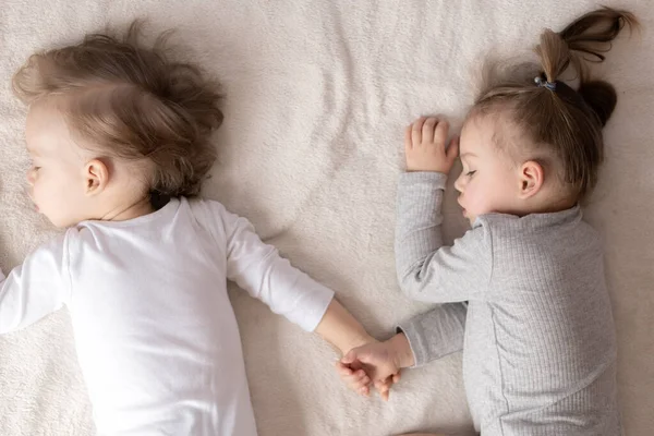 Barndom, sömn, avkoppling, familj, livsstil koncept - två små barn 2 och 3 år gammal klädd i vit och beige body suit sova på en beige och vit säng vid lunch håller händerna ovanifrån. — Stockfoto