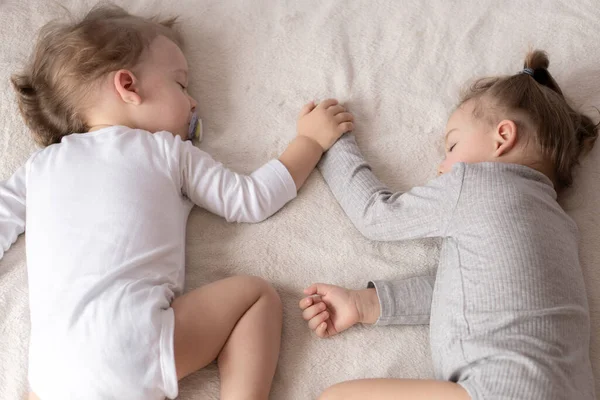 Enfance, sommeil, détente, famille, mode de vie concept - deux jeunes enfants de 2 et 3 ans habillés en body blanc et beige dorment sur un lit beige et blanc au déjeuner en se tenant la main vue du dessus . — Photo