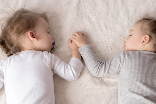 Infância, sono, relaxamento, família, conceito de estilo de vida - duas crianças de 2 e 3 anos de idade vestidas com um bodysuit branco e bege dormem em uma cama bege e branca no almoço de mãos dadas vista superior . — Fotografia de Stock