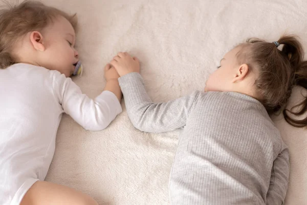 Infancia, sueño, relajación, familia, concepto de estilo de vida: dos niños pequeños de 2 y 3 años vestidos con traje de cuerpo blanco y beige duermen en una cama beige y blanca en el almuerzo tomados de la mano vista superior . — Foto de Stock