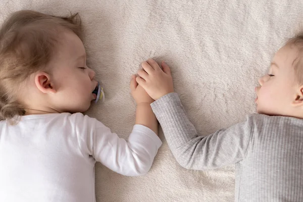 Barndom, sömn, avkoppling, familj, livsstil koncept - två små barn 2 och 3 år gammal klädd i vit och beige body suit sova på en beige och vit säng vid lunch håller händerna ovanifrån. — Stockfoto