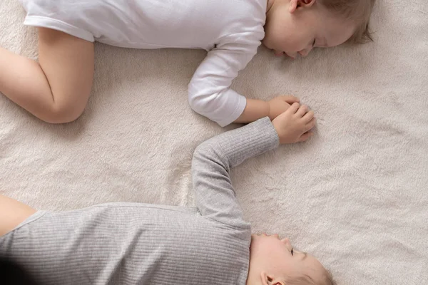 Infancia, sueño, relajación, familia, concepto de estilo de vida: dos niños pequeños de 2 y 3 años vestidos con traje de cuerpo blanco y beige duermen en una cama beige y blanca en el almuerzo tomados de la mano vista superior . — Foto de Stock