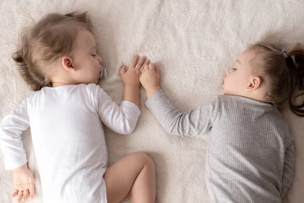 Infancia, sueño, relajación, familia, concepto de estilo de vida: dos niños pequeños de 2 y 3 años vestidos con traje de cuerpo blanco y beige duermen en una cama beige y blanca en el almuerzo tomados de la mano vista superior . — Foto de Stock