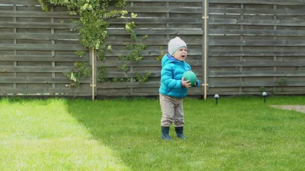 Infância, lazer, jogo, quintal, conceito de primavera - três crianças pequenas brincam no quintal com bolas e um fitball em uma grama verde no início da primavera em tempo frio . — Vídeo de Stock