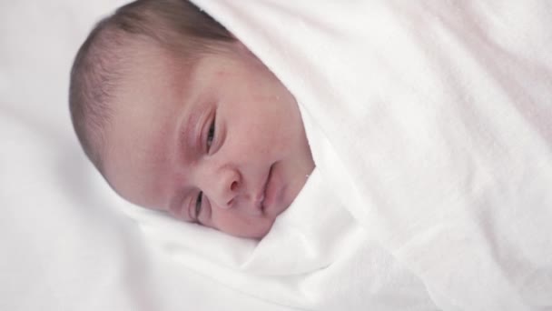 Kindertijd, kindertijd, ouderschap, moederschap concept - close-up portret van een pasgeboren baby liggend op een wit beige bed gewikkeld in een luier ontwaakt wakker opent zijn ogen en steekt zijn tong uit. — Stockvideo