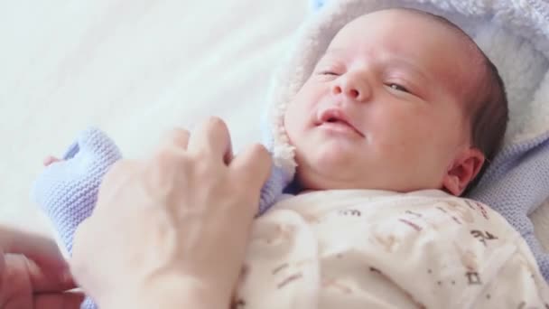 Παιδική ηλικία, βρεφική ηλικία, μητρότητα, οικογενειακή έννοια - γκρο πλαν ενός γυναικείου χεριού βάζοντας ένα νεογέννητο μωρό σε μια μπλε μπλούζα σε ένα μπεζ κρεβάτι σε λευκό φόντο. Η μαμά ντύνει το μωρό.. — Αρχείο Βίντεο