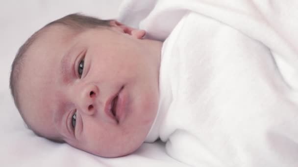 Infancia, infancia, paternidad, concepto de maternidad - retrato de cerca de un bebé recién nacido acostado en una cama de color beige blanco envuelto en un pañal despierto cortando los ojos y sacando la lengua . — Vídeos de Stock