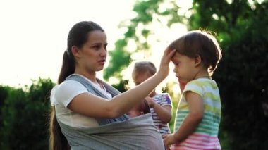 aile, çocuklar, annelik, eğlence ve insan konsepti. Parkta gün batımının arka planında aynı yaşta iki büyük çocukla takılan askılı bebeğiyle pek çok çocuğun genç annesi..