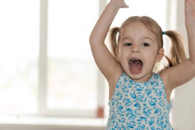Çocukluk, insanlar, duygu konsepti - iki at kuyruklu ve mavi elbiseli küçük kız ellerini kaldırır ve pencereye doğru bağırır.