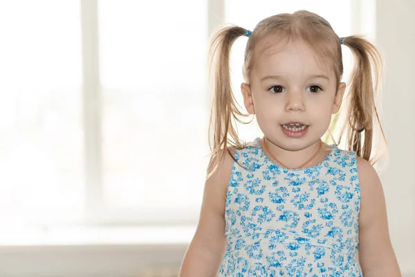 Infância, pessoas, conceito de emoções - menina de plano médio com dois rabos de cavalo e um vestido azul sorri e mostra emoções no fundo da janela — Fotografia de Stock