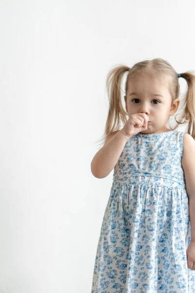 Kindheit, emotionales Konzept - mittlerer Plan isolieren kleine süße dreijährige Mädchen mit zwei Pferdeschwänzen in einem blauen Kleid bläst in eine Faust und zeigt Fratzen auf weißem Hintergrund. — Stockfoto
