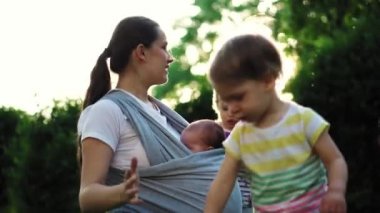 aile, çocuklar, annelik, eğlence ve insan konsepti. Parkta gün batımının arka planında aynı yaşta iki büyük çocukla takılan askılı bebeğiyle pek çok çocuğun genç annesi..