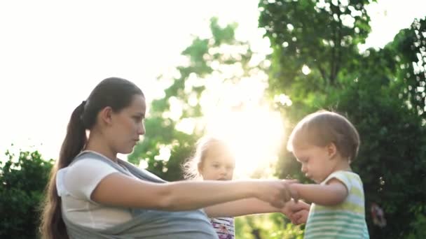 Família, crianças, maternidade, entretenimento e conceito de pessoas - jovem mãe bonito de muitas crianças com seu bebê na funda brinca com duas crianças mais velhas da mesma idade no fundo do pôr do sol no parque . — Vídeo de Stock