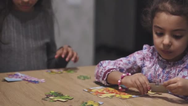 La infancia, los juegos, el ocio, el aislamiento y el concepto de aprendizaje a distancia: dos hermanas persas de pelo negro y pelo negro de aspecto de Oriente Medio, adolescentes y niños, ponen puzzles sobre la mesa en casa.. — Vídeo de stock