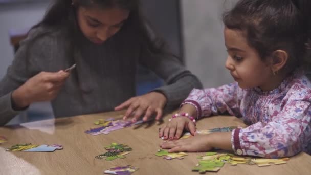 Barndom, spel, fritid, isolering och distansundervisning koncept - två mörkhåriga svarthåriga persiska flickor systrar i Mellanöstern utseende, tonåring och barn lägger pussel på bordet hemma. — Stockvideo