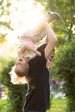 Babalık, ebeveynlik, çocukluk, şefkat, yaz ve eğlence konsepti - sakallı ve siyah tişörtlü genç baba kollarında küçük oğlunu parktaki günbatımının arka ışığında tutuyor..