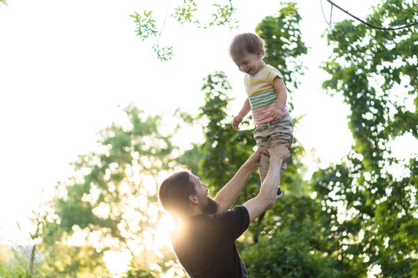 Fatherhood, parenthood, childhood, caring, summer and leisure concept - young dad with beard and long hair in black t-shirt raises up his son in his arms in the backlight of the sunset in the park. — Stock Photo, Image