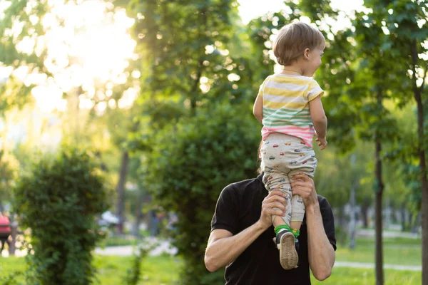 Apaság, szülőség, gyermekkor, gondoskodás, nyár és szabadidő koncepció - fiatal apa szakállal és hosszú haj fekete pólóban tartja a karját kisfia a háttérben a naplemente a parkban. — Stock Fotó