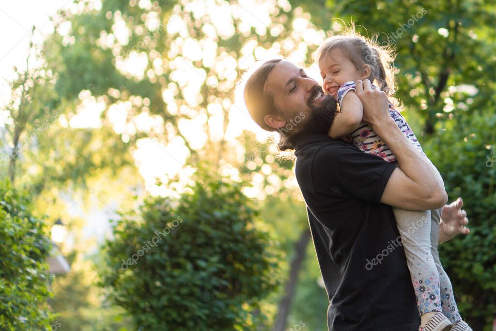 Fatherhood, parenthood, childhood, caring, summer and leisure concept - young dad with beard and long hair in black t-shirt holding his little daughter in his arms in backlight of sunset in the park.