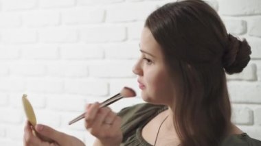 Moda ve güzellik, partiler, kozmetoloji, yaşam tarzı konsepti - makyaj yapan genç, koyu renkli, sıradan bir kız yanaklarını kahverengileştiriyor ve beyaz tuğladan bir arka planda sevimli gülümsüyor.
