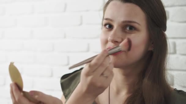 Mode en schoonheid, feesten, cosmetologie, life style concept - jong mooi donker getint casual meisje doen make-up bruint haar wangen en glimlacht schattig op een witte bakstenen achtergrond. — Stockvideo