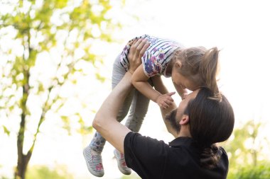 Babalık, ebeveynlik, çocukluk, şefkat, yaz ve eğlence konsepti - sakallı ve siyah tişörtlü genç baba, parkta gün batımının ışığında küçük kızını kollarında tutuyor..