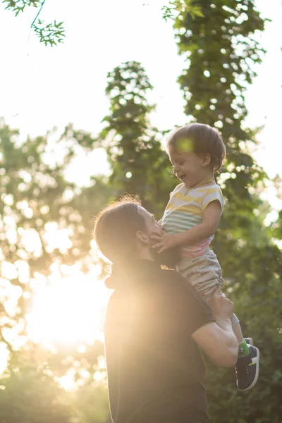 Apaság, szülőség, gyermekkor, gondoskodás, nyár és szabadidő koncepció - fiatal apa szakállal és hosszú haj fekete pólóban tartja a karját kisfia a háttérben a naplemente a parkban. — Stock Fotó