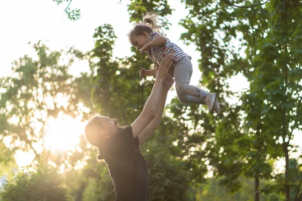 Apaság, szülőség, gyermekkor, gondoskodás, nyár és szabadidő koncepció - fiatal apa szakállal és hosszú haj fekete pólóban feldobja kislányát a háttérben a naplemente a parkban. — Stock Fotó