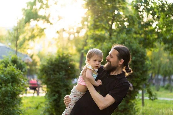 Fatherhood, parenthood, childhood, caring, summer and leisure concept - young dad with beard and long hair in black t-shirt holds in his arms little son in the backlight of the sunset in the park. — Stock Photo, Image