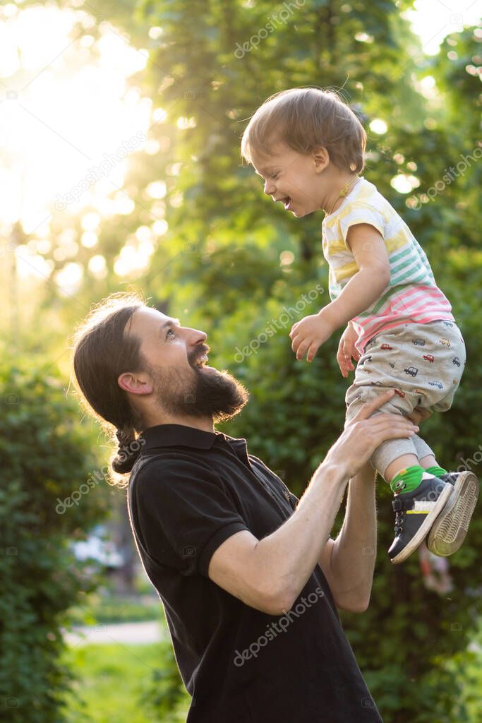 Fatherhood, parenthood, childhood, caring, summer and leisure concept - young dad with beard and long hair in black t-shirt holds in his arms little son in the backlight of the sunset in the park.
