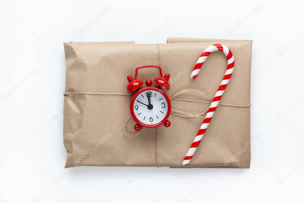 Christmas gift wrapped in brown craft paper, tied with scourge, decorated with cane candy and small analog clock on white background. Minimal style. Top view. Do it yourself, celebration, eco concept