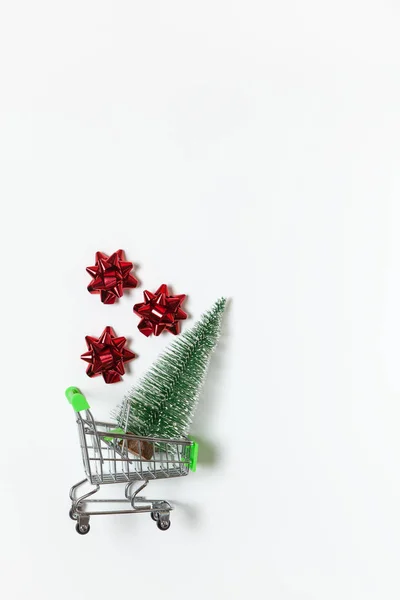 Abete di Natale e fiocchi rossi per regalo nel carrello della spesa giocattolo su sfondo bianco, copiare lo spazio. Capodanno, vendite, shopping online. Verticale, piatta. Stile minimale. Vista dall'alto. Per i social media — Foto Stock