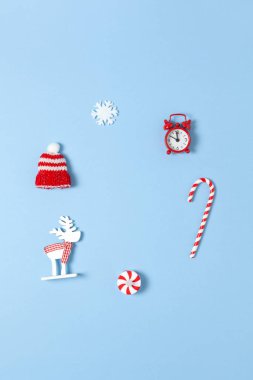 Basit yaratıcı Noel kompozisyonu. Şeker kamışı, geyik, şapka, kar tanesi, mavi arka planda kırmızı saat, kopyalama alanı. Asgari düzeyde. Üst Manzara. Dikey. Sosyal medya, tebrik kartı, ofis malzemeleri