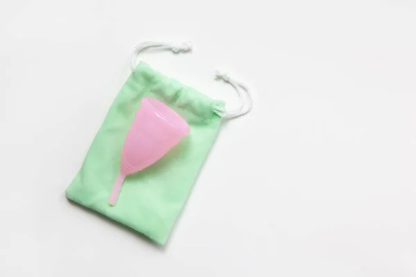 Taza menstrual rosa sobre bolsita verde sobre fondo blanco. Concepto cero desperdicio, ahorros, minimalismo, en estos días. Producto de higiene femenina, plano, espacio de copia. Horizontal — Foto de Stock