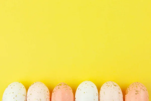 Creatively painted eggs in pastel colors decorated with gold leaf are arranged in row along bottom edge on yellow background, copy space. Happy Easter DIY concept. Flat lay. Horizontal