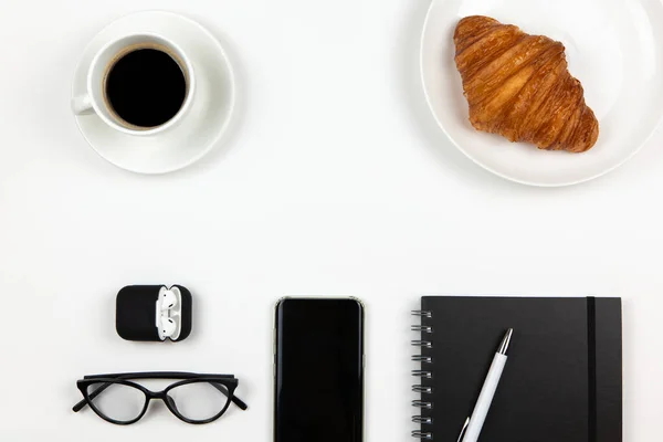 Modern working space of freelancer. Smart phone, coffee, notebook, pen, croissant, glasses, headphones in case on white background, copy space, flat lay. Business breakfast, work from home concept.