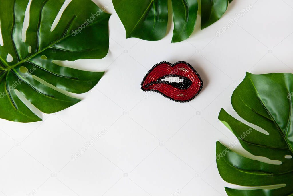 Fashionable brooch in shape of lips from Japanese beads, green leaves of Monstera on white background. Close-up, flat lay, copy space. Concept trends, fashion, style, DIY, needlework