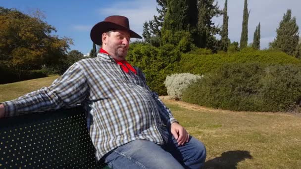 Cowboy tips his brown hat and shines sitting on bench in city park — Stock Video