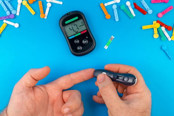 Test na cukrzycę. Ręce mężczyzny sprawdzają poziom cukru we krwi za pomocą miernika glukozy na niebieskim tle. Urządzenie do pomiaru poziomu cukru we krwi. Paski testowe, tabletki na niebieskim tle. — Zdjęcie stockowe
