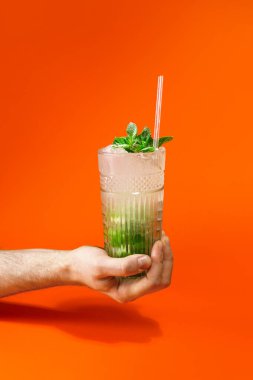 Turuncu arka planda bir bardak tropikal mojito kokteyli tutan bir adamın eline yakın çekim. Yaz tatili konsepti.