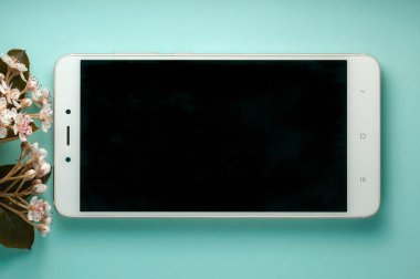 Mavi yüzey üzerinde çiçek açan dalların yakınında boş ekran ile beyaz akıllı telefonun üst görünümü