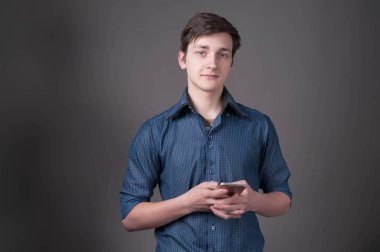 Gömleğinin kolları sarılı, akıllı telefon tutan ve gri arka planda kameraya bakan kendine güvenen yakışıklı bir genç.