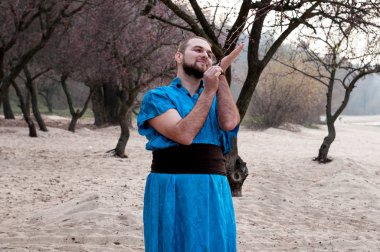 Mavi kimonolu yakışıklı, kurnaz ve sakallı adam. Siyah kuşaklı, gülümseyen, kumdaki ağacın dalını tutan.