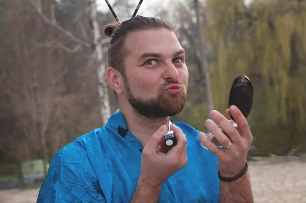 handsome sly man with beard and hair bun in blue kimono looking at camera, holding pocket mirror and applying red lipstick in front of tree in park