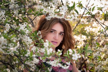 Ağaçların arkasında çiçek açan kiraz dallarıyla duran ve bahçedeki kameraya bakan uzun saçlı güzel kadının yumuşak odağı.