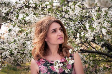 Çiçek desenli uzun saçlı güzel kadının seçici odağı bahçesinde beyaz çiçeklerle çiçek açan kiraz ağacının önünden uzağa bakıyor.